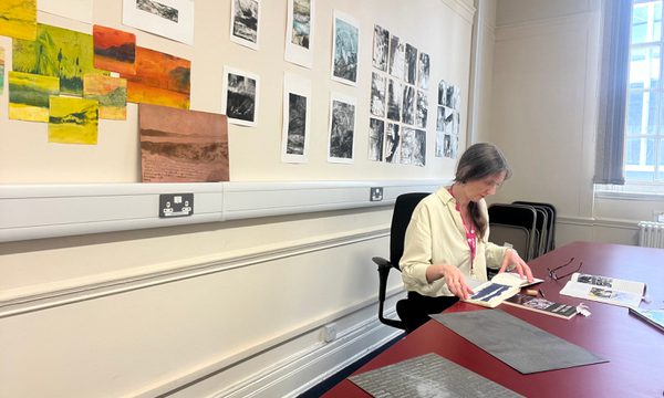 Fae in her studio space sitting at a desk with artworks on the wall behind.