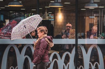 A woman walks by with an umbrella shielding her from the rain. Behind her is a cafe and you can see people seated inside at the window.