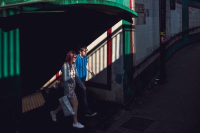 A man and a woman emerge from a dark underpass into the sunshine. They are both wearing sunglasses.