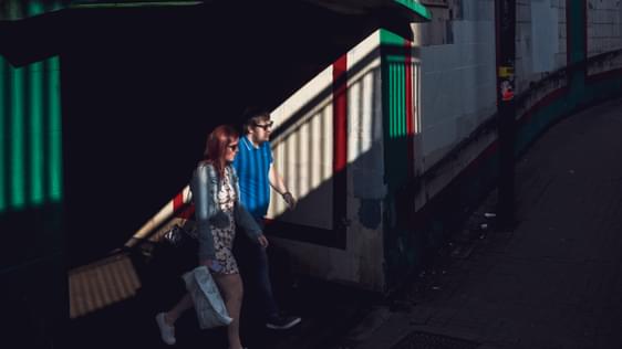 A man and a woman emerge from a dark underpass into the sunshine. They are both wearing sunglasses.