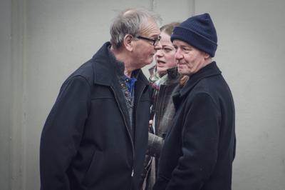 Two men are standing close to each other, and a woman is close by behind. The men are looking in different directions and not speaking. It is unclear what is happening.