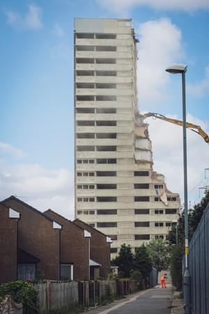 In the background a tower block is being demolished, and in the foreground you can see a workman sweeping the streets outside a row of houses.