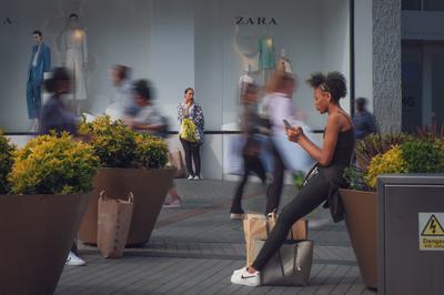 A young woman is looking at her phone, and further back there is a woman who appears to be standing and waiting. In between them are the blurred figures of people walking in the busy area of the Bullring.