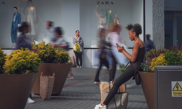 A young woman is looking at her phone, and further back there is a woman who appears to be standing and waiting. In between them are the blurred figures of people walking in the busy area of the Bullring.