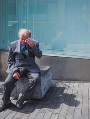 An elderly man is sitting on a concrete block, holding a hankerchief to one of his eyes. He is wearing a grey jacket, trousers and trainers, a blue short and a green tie.