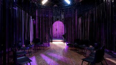 A dark room with empty chairs, a purple light shines on entrance curtain.