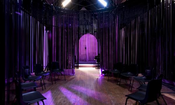 A dark room with empty chairs, a purple light shines on entrance curtain.