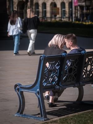 A couple sitting on a park bench, they are turnbed to each other and their foreheads and noses are touching.