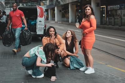 Four women are in the street in the city centre, three are on the floor and one is standing. One is holding a bottle of wine and another is holding a full glass.