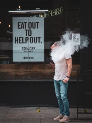 A man is vaping and the white smoke covers his upper body and face. He seems to be staring at his phone through the white smoke. Behind him is a sign that says: Eat out to help out.