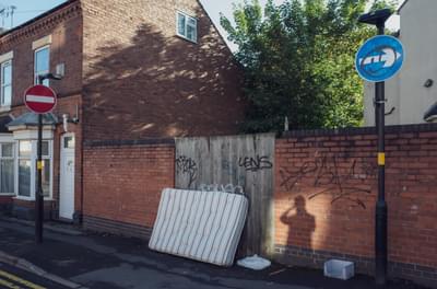 An abandoned mattress is propped up against a gate and wall in a street of terraced houses. The shadow of the photographer is captured on the wall next to the mattreess.