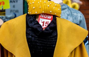 A shop dummy wearing a yellow coat. The dummy doesn't have a head, but there is a soft toy that looks like a yellow smiling emoji on the wall behind it, and this looks like the dummy's head.