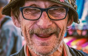 A smiling man wearing a hat covered in a variety of colourful badges. He is wearing glasses and has a greying beard.
