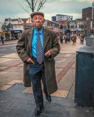 An elderly man is walking in the street. He is wearing a bright blue shirt and a patterned tie, pin striped trousers, and a tweed overcoat and cap.