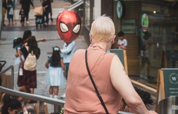 A woman can be seen from the back looking down at the street below. A ballow of Spiderman's face rises up from below and seems to stare in her face.