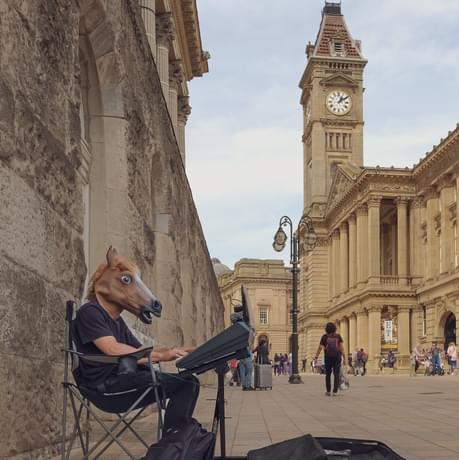 A person wearing a horse mask is busking in the street. Birmingham Museum and Art Gallery can be seen in the background.