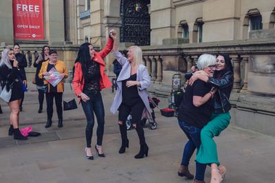 Women dance and embrace in the street while others look on smiling.