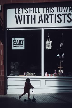 The silhouette of a small child on a scooter can be see in front of a shop front. Above the shop window it says: Let's fill this town with artists.