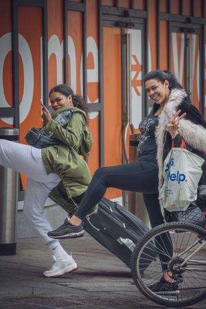 Two women stand in a similar high step pose. They are smiling. One can be seen pulling a suitcase behind her. They appear to be outside the train station.