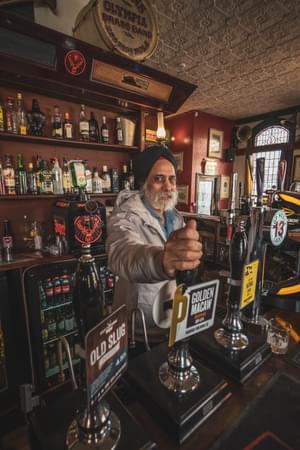 A grey bearded man in a turban stands behind the bar of a pub.