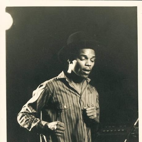 Ranking Roger of The Beat. A black man wearing a stripy shirt and  a black hat. He looks to be mid dance movement.