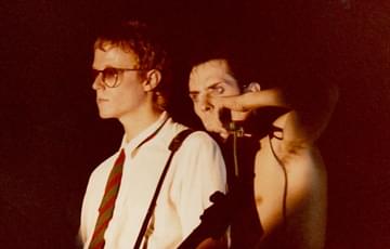 David J and Pete Murphy of Bauhaus. A man stand in front of another. The man in front has a white shirt, tie, sunglasses and play the guitar. Behind him a topless man looks menacingly out to the crowd whilst singing into a mic