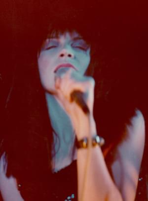 Annie Lennox of Eurythmics. The image is fuzzy, slightly out of focus. Annie has long bushy dark hair and pink lipstick. She has her eyes closed as she sings into the mic.