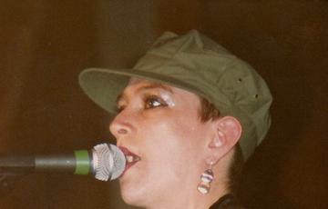 Lesley Woods of Au Pairs.She wears a black top and a green hat. She's got short hair, glittery eye makeup and dark lipstick. She's playing the guitar and singing into a mic