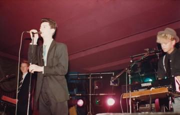 Three members of band Depeche Mode on stage. The frontman is wearing a dark grey suit, white shirt and blue tie. The keyboard player is also in a suite and tie. The Keys player looks more casual in a black shirt, scruffy hair and a hat