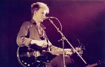 Edwyn Collins of Orange Juice. They are singing into a mic, have short hair and a guitar.
