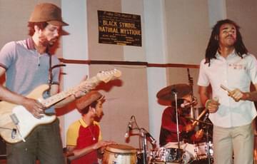Handsworth reggae band Natural Mystique playing a show at Birmingham University’s High Hall. There are four black men on stage. A guitarist, two drummers and a front man.