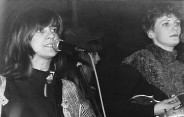 Two members of band called The Raincoats. Both are female. The one on the left has long dark hair and sings into a mic. The woman on the right has short hair and plays a guitar
