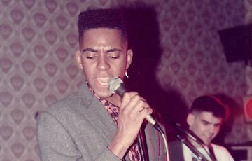 Colbert Hamilton AKA Black Elvis. He's singing into a mic. He wears a pink leopard print shirt and a grey blazer. He has a neat box hair cut. In the distance a white man plays a red guitar.