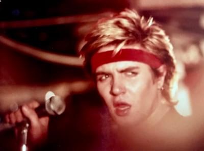 Simon Le Bon of Duran Duran . He's holding a mic, and looks to the right of the camera. It's a close portrait. He's got blonde spiky hair and a red headband