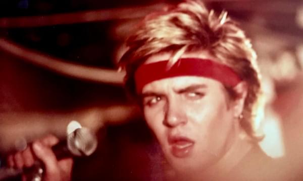 Simon Le Bon of Duran Duran . He's holding a mic, and looks to the right of the camera. It's a close portrait. He's got blonde spiky hair and a red headband