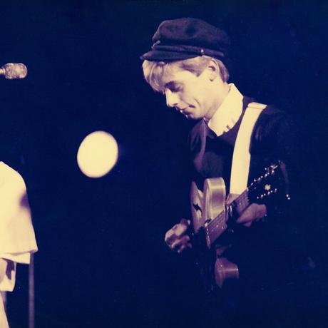 Two Haircut 100 band members on stage. Both hold guitars, a mic stands between them. the two members are male, young, with short blonde hair. One wears a white shirt and scarf, the other wears a black hat, black jacket and white shirt. n the distance a sax player is just visible.