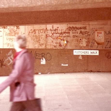 A concrete underpass covered in graffiti. A person is walking past the camera, they're blurry and looking away form the camera, towards the other side of the underpass