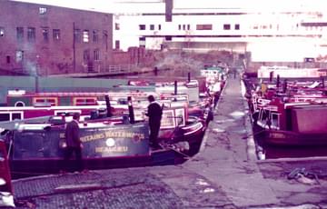 Narrowboats moored in wide canal in Birmingham city centre. Down the centre is a jetty, with boats more either side. In the distance there's a white factory building