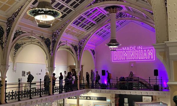 A large vaulted gallery space with ironwork arches, balcony around the edge of the first floor with people on the ground floor gallery below, with a large neon sign at the bottom.