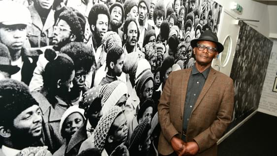 Vanley Burke stands in front of an image from one of his photographs that has been blown up to be very large in size.
