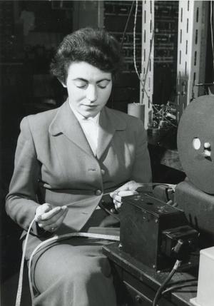 Black and white photo of a lady (Steve Shirley) sitting working with computer machinery to the side and behind.