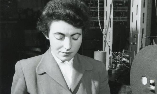 Black and white photo of a lady (Steve Shirley) sitting working with computer machinery to the side and behind.
