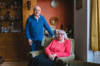 Tony is standing next to Alicja who is sitting in an armchair. They are in her living room.