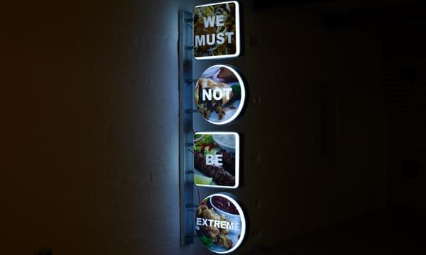 There is a vertical neon light in four parts. The words in the lights read We Must Not Be Extreme.