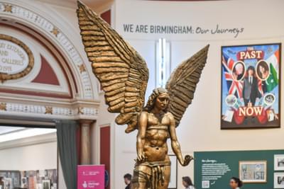 The bronze statue of the archangel Lucifer. The male statue is naked. Lucifer has a feminine face and long hair. He has large wings that point upwards. His hands are outstretched and the palms of his hands are facing down.