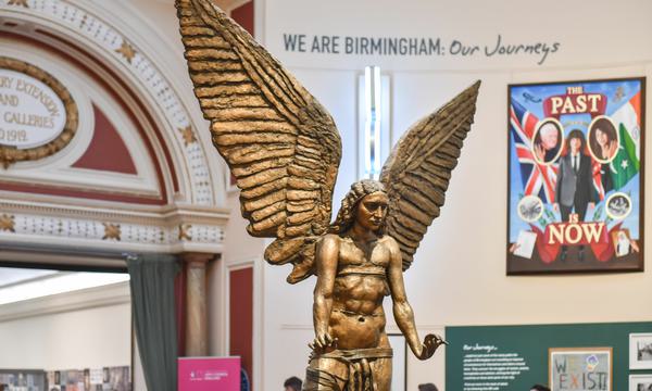 The bronze statue of the archangel Lucifer. The male statue is naked. Lucifer has a feminine face and long hair. He has large wings that point upwards. His hands are outstretched and the palms of his hands are facing down.