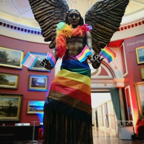The statue of Lucifer is wearing a rainbow coloured feather boa and a rainbow coloured wrap around that is covering his body. He is holding 2 small light blue, pink and white Transgender flags in one hand and 2 small rainbow LGBTQ flags in the other hand.