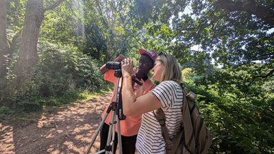Two people surrounded by trees are looking into a camera on a tripod.