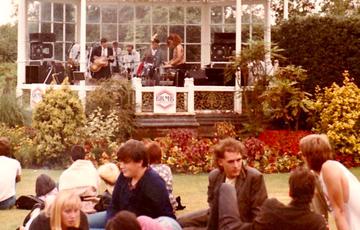 A group of young men and women are sitting and lying on the grass. Behind them a band can be seen playing in a white bandstand.
