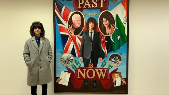 A young woman artist standing next to her artwork The Past is Now.  She has long dark hair and is wearing a grey overcoat. She is the woman portrayed in the centre of the artwork.